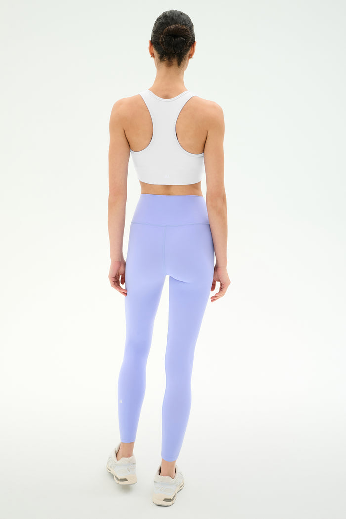 A woman faces away, wearing the SPLITS59 Airweight Bra with Ruching in white, paired with light purple leggings and white sneakers, set against a plain white background.