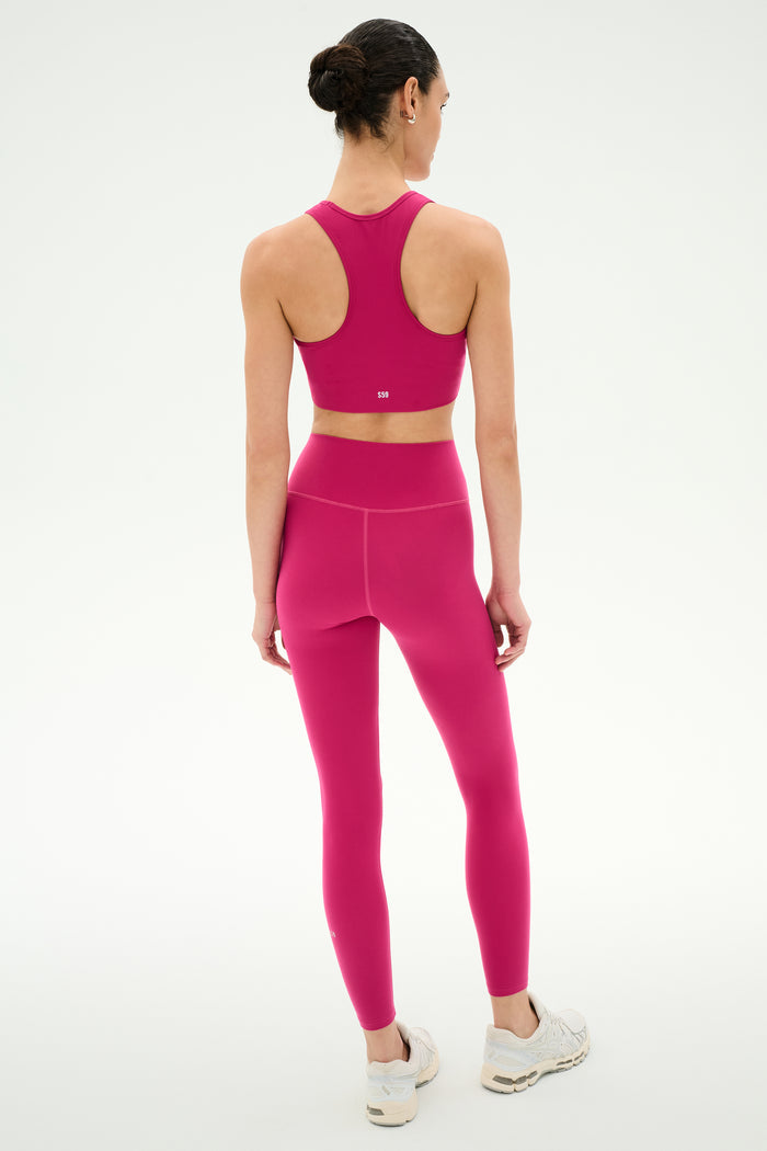 A person stands facing away, dressed in the SPLITS59 Airweight High Waist Legging in Sangria with a coordinating pink sports bra and white sneakers—an ideal look for hot yoga sessions—set against a plain white background.