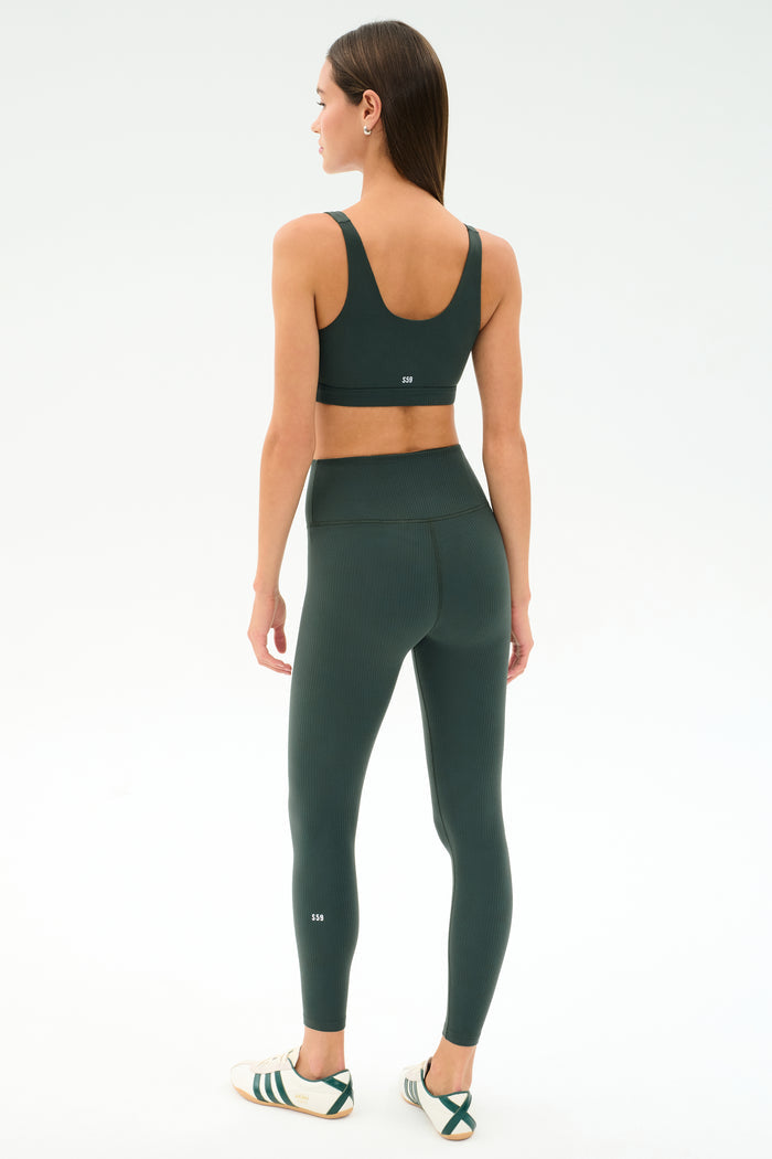 A woman faces away, wearing the SPLITS59 Airweight Rib Bra - Military, matching high-waisted leggings, and white sneakers with green stripes against a plain white background.