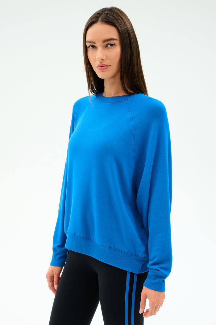 A woman stands against a plain background, wearing the SPLITS59 Andie Oversize Fleece Sweatshirt in Classic Blue and black leggings with blue stripes, facing slightly to the side.