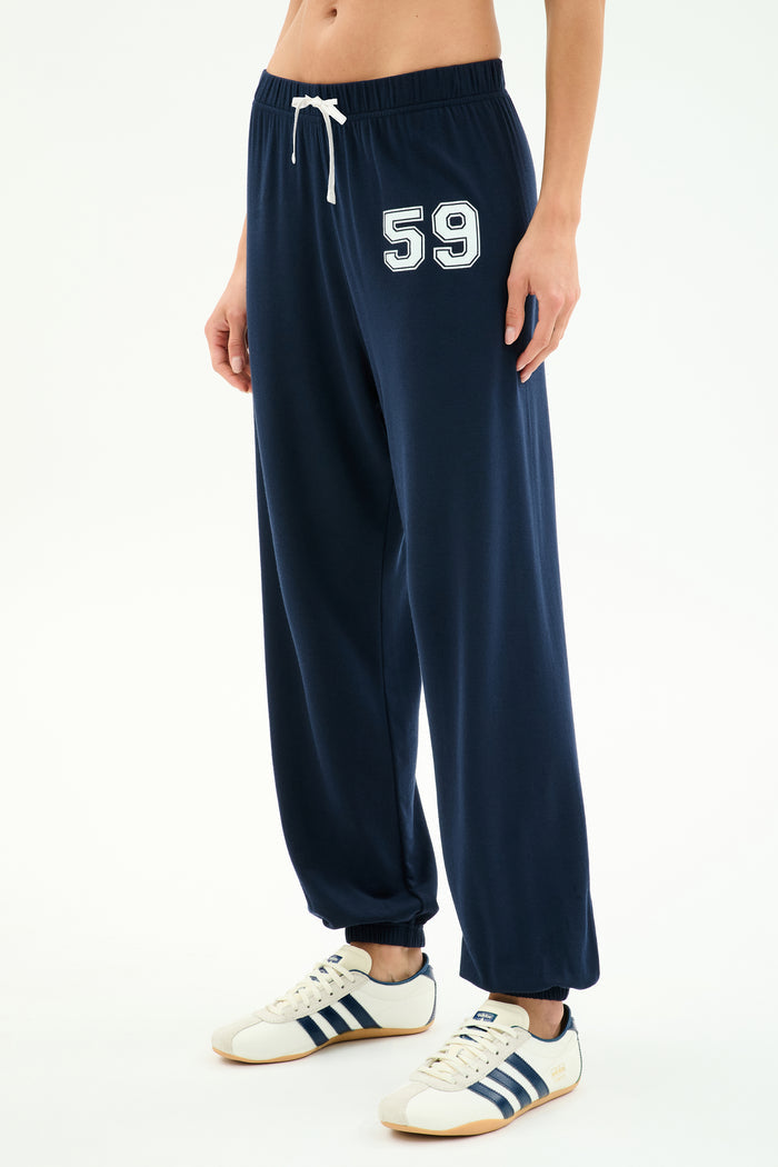 A person wears the SPLITS59 Andie Oversized Fleece Varsity 59 Sweatpant in Indigo/59, paired with white sneakers with navy stripes, standing against a white background. Made in Los Angeles.