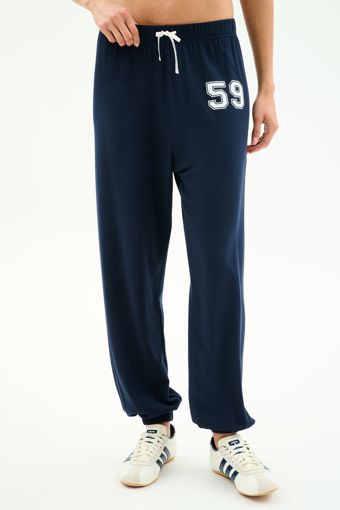 Wearing SPLITS59 Andie Oversized Fleece Varsity 59 Sweatpant in Indigo with a white drawstring and "59" on the left thigh, paired with white sneakers featuring navy stripes. Made in Los Angeles for superior comfort and style.