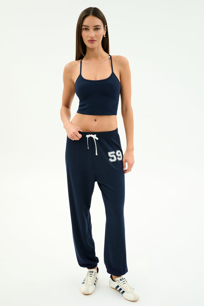 A woman stands against a plain white background, wearing a navy crop top, white sneakers, and SPLITS59 Andie Oversized Fleece Varsity 59 Sweatpant in Indigo/59 with “59” on the left leg.