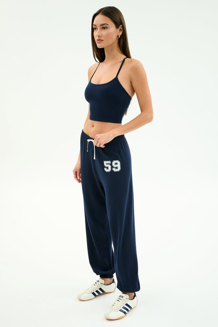 A woman wears the SPLITS59 Andie Oversized Fleece Varsity 59 Sweatpant in Indigo/59, paired with a navy crop top and white sneakers with navy stripes, standing against a plain white background. Made in Los Angeles.