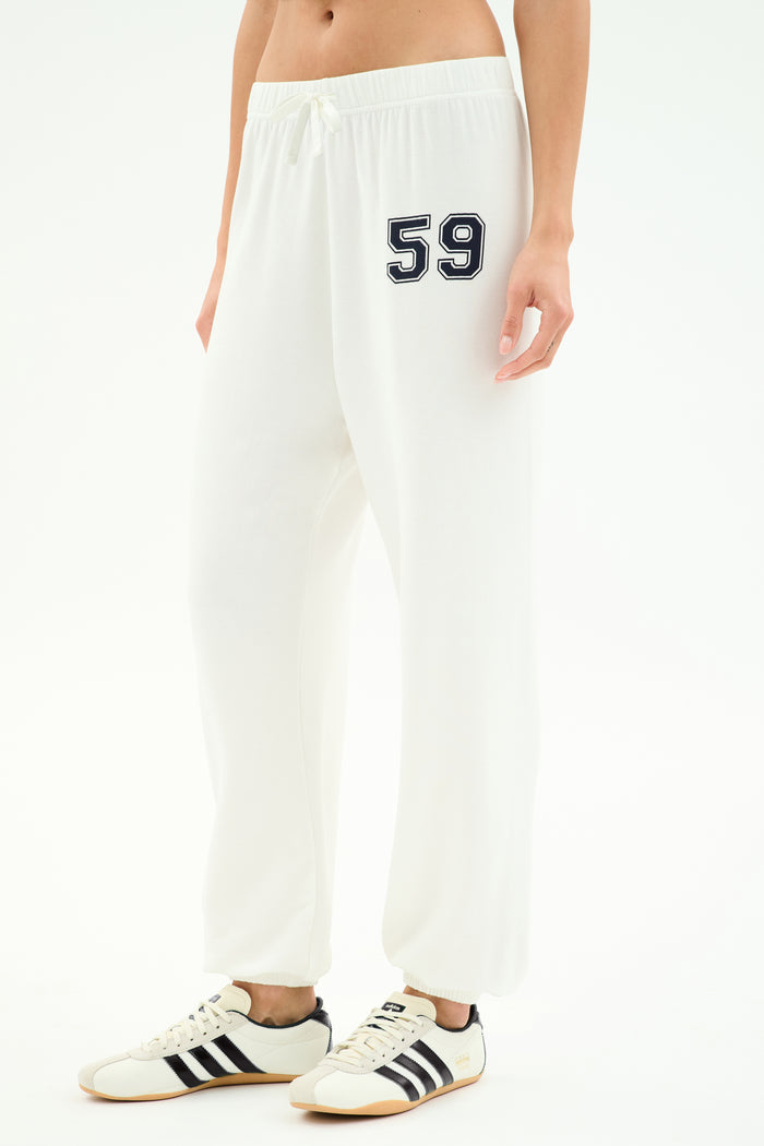 A person in SPLITS59 Andie Oversized Fleece Varsity 59 Sweatpant—white with bold "59"—paired with white sneakers with black stripes, perfect for cool weather workouts. Made in Los Angeles.