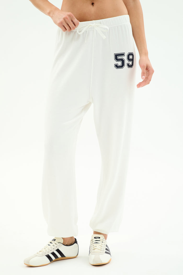 A person wears SPLITS59's Andie Oversized Fleece Varsity 59 Sweatpant in White/59, featuring the number 59 on the left hip, paired with white sneakers with black stripes—ideal for cool weather workouts—against a plain white background.