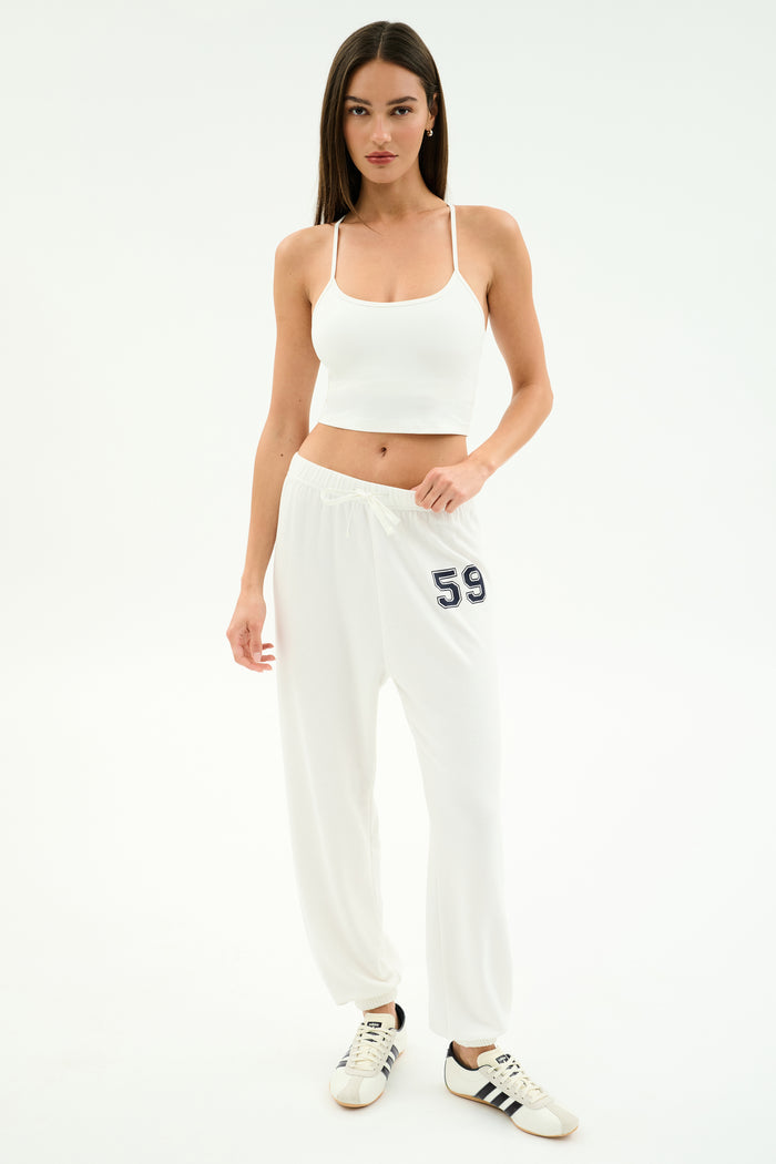Woman standing against a white background wears a white crop top, SPLITS59 Andie Oversized Fleece Varsity 59 Sweatpant in White/59, and white Adidas sneakers—ideal for cool weather workouts. MADE IN LOS ANGELES.