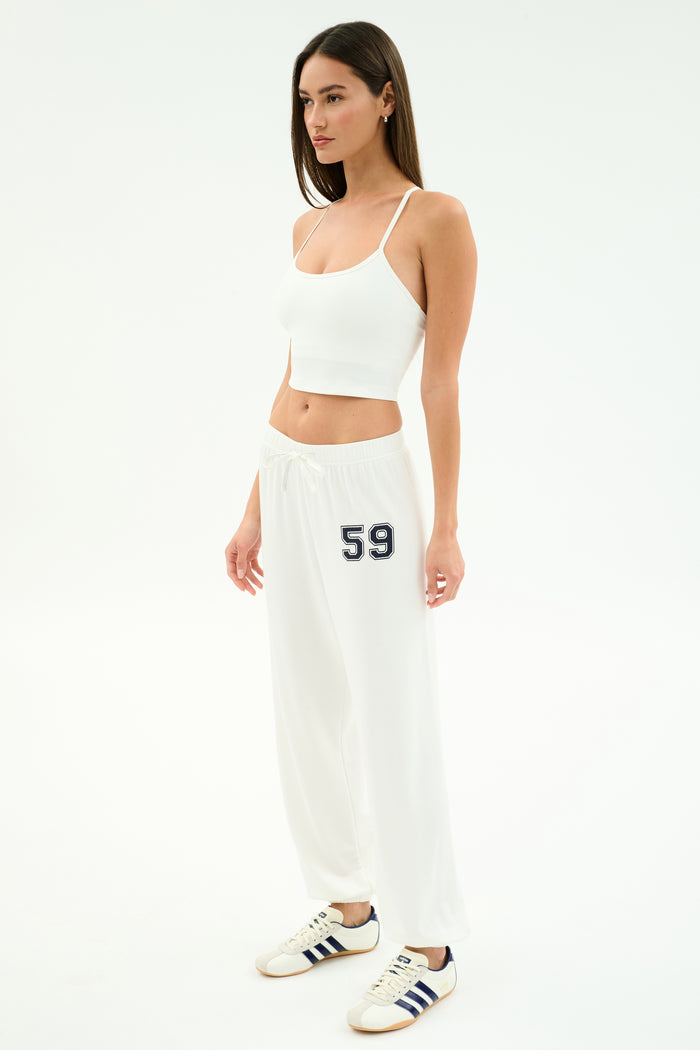 A woman in a white studio wears a white crop top and the SPLITS59 Andie Oversized Fleece Varsity 59 Sweatpant (White/59), ideal for cool weather workouts, finished with white sneakers with navy stripes.