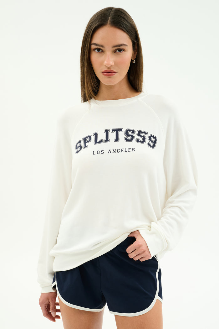 A woman wears the SPLITS59 Andie Oversized Fleece Varsity Graphic Sweatshirt in white with "SPLITS59 LOS ANGELES" print and navy athletic shorts with white trim, standing against a plain light background.