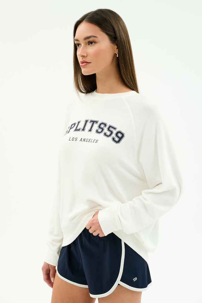 A woman wears a white SPLITS59 Andie Oversized Fleece Varsity Graphic Sweatshirt with "Splits59 Los Angeles" and navy athletic shorts, standing against a plain white background.