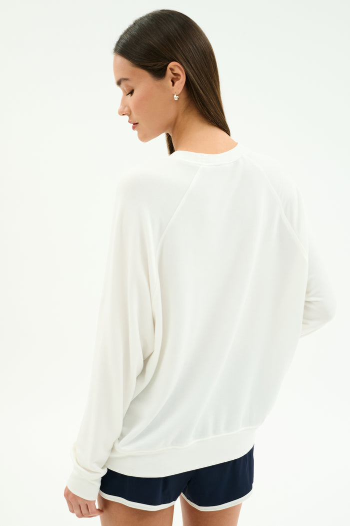 A woman faces away, wearing the SPLITS59 Andie Oversized Fleece Varsity Graphic Sweatshirt in white against a plain background. This crewneck pullover is made in Los Angeles.