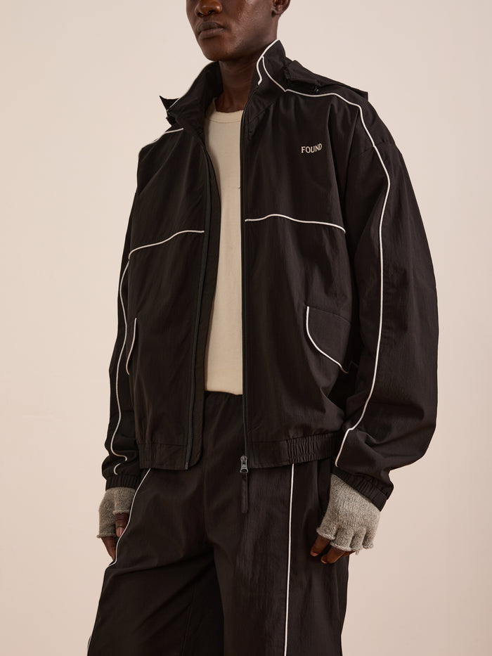 Someone stands against a plain background wearing the FOUND Arena Stripe Track Jacket, a unisex black track jacket with white piping, paired with matching pants, a beige shirt, and gray fingerless gloves. The set is made from lightweight technical fabric.