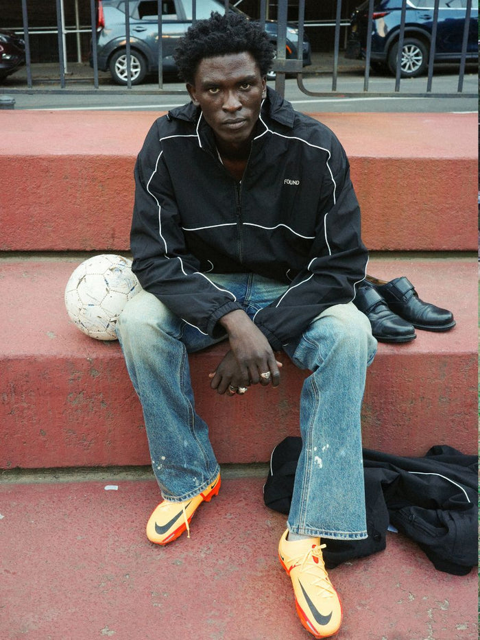 A man sits on red steps with a soccer ball, wearing FOUND’s Arena Stripe Track Jacket, jeans, and bright orange cleats; dress shoes and a black garment rest on the step.