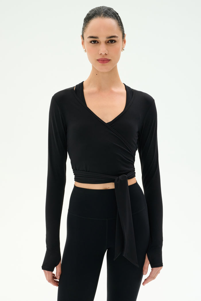 A woman wears the SPLITS59 Bella Airweight Lite Wrap in black—a long-sleeve, wrap-style crop top—paired with high-waisted black leggings, posed against a plain white background.