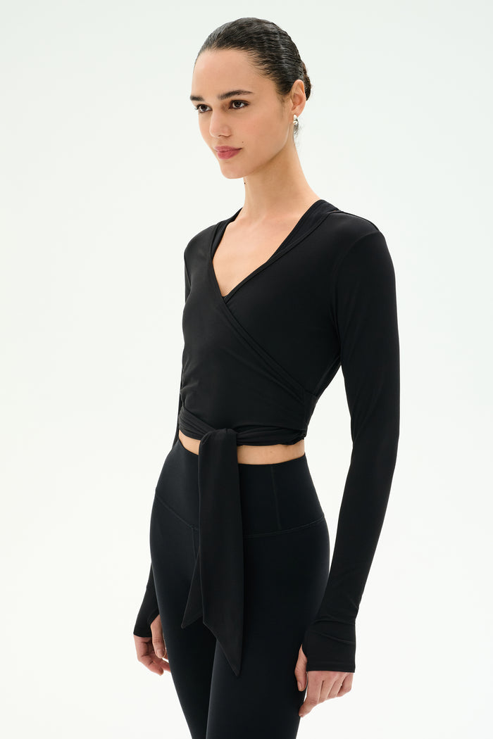A woman stands against a plain white background wearing the SPLITS59 Bella Airweight Lite Wrap in black, a stylish long-sleeve spandex top with a front tie, paired with black high-waisted leggings.