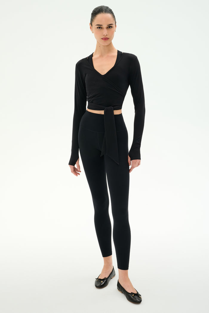 A woman stands against a plain white background wearing the SPLITS59 Bella Airweight Lite Wrap in black, paired with black leggings and black ballet flats.