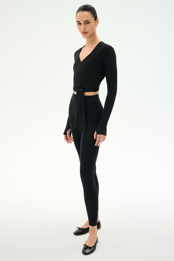 A person stands against a white background wearing the SPLITS59 Bella Airweight Lite Wrap in black, paired with black spandex leggings and black ballet flats.