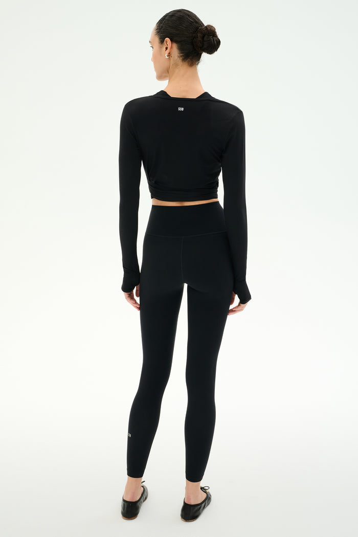 A person faces away, wearing the SPLITS59 Bella Airweight Lite Wrap in black and black spandex leggings with ballet flats, set against a plain light background.