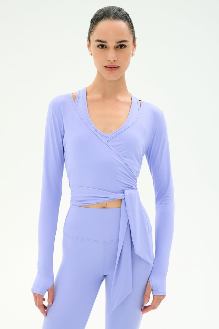 A woman wearing the SPLITS59 Bella Airweight Lite Wrap top in Purple Haze with matching high-waisted leggings stands against a plain white background.