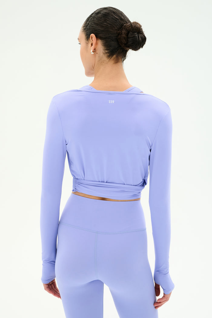 A woman stands with her back to the camera, wearing the SPLITS59 Bella Airweight Lite Wrap in Purple Haze, paired with matching leggings. Her hair is styled in a neat bun against a plain light background.