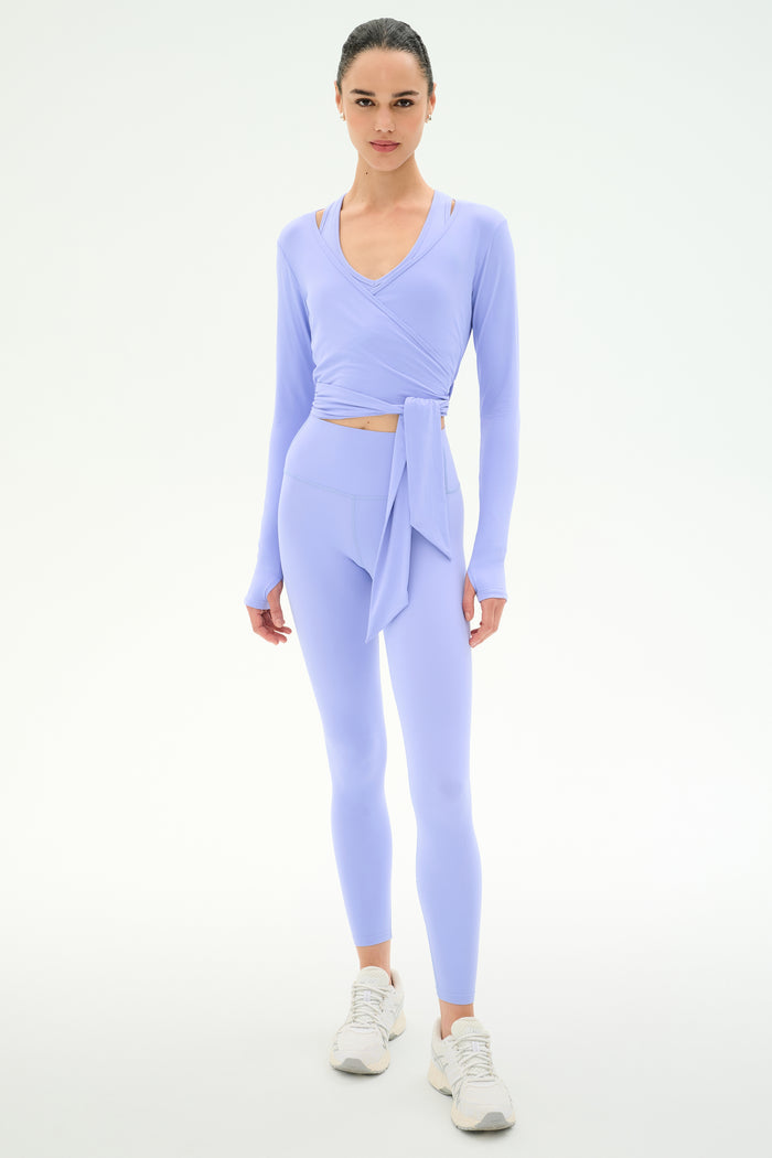 A woman faces forward against a white background, wearing the SPLITS59 Bella Airweight Lite Wrap in Purple Haze, paired with matching leggings and white sneakers.