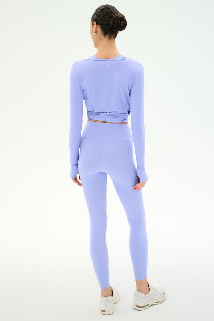 A woman faces away, wearing the SPLITS59 Bella Airweight Lite Wrap in Purple Haze—a light purple nylon-spandex set—paired with white athletic shoes, against a plain light background.