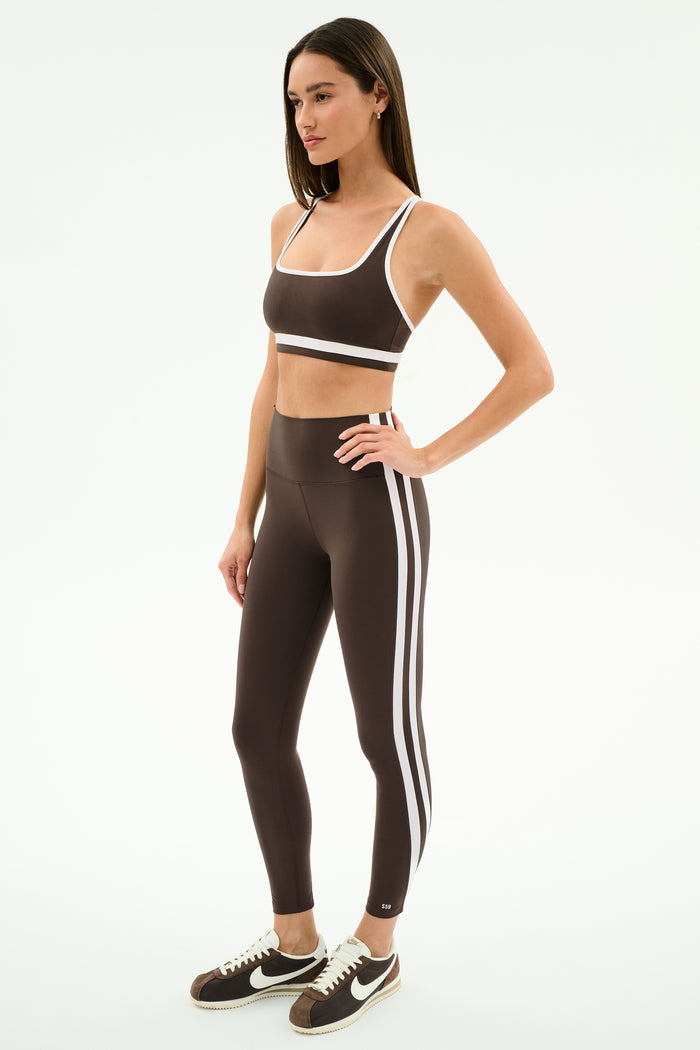 A woman wears the SPLITS59 Bianca Rigor Bra in Dark Chocolate/White and matching high-waisted leggings with white stripes, paired with black and white Nike sneakers—an ideal gym outfit—against a plain white background.