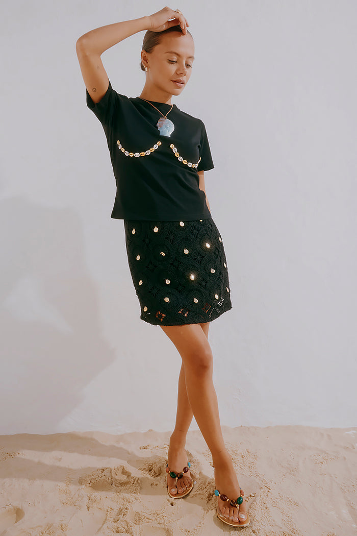 Black crochet wrap skirt with gold shell embellishments, styled with a shell-detail t-shirt on sand.