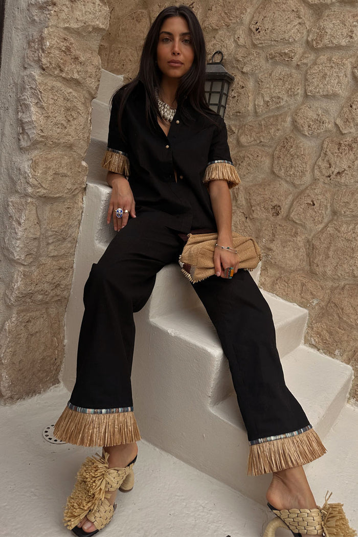 Black Raffia Marra Trousers with a raffia fringe hem, styled as a co-ord on a model sitting on white stone stairs.