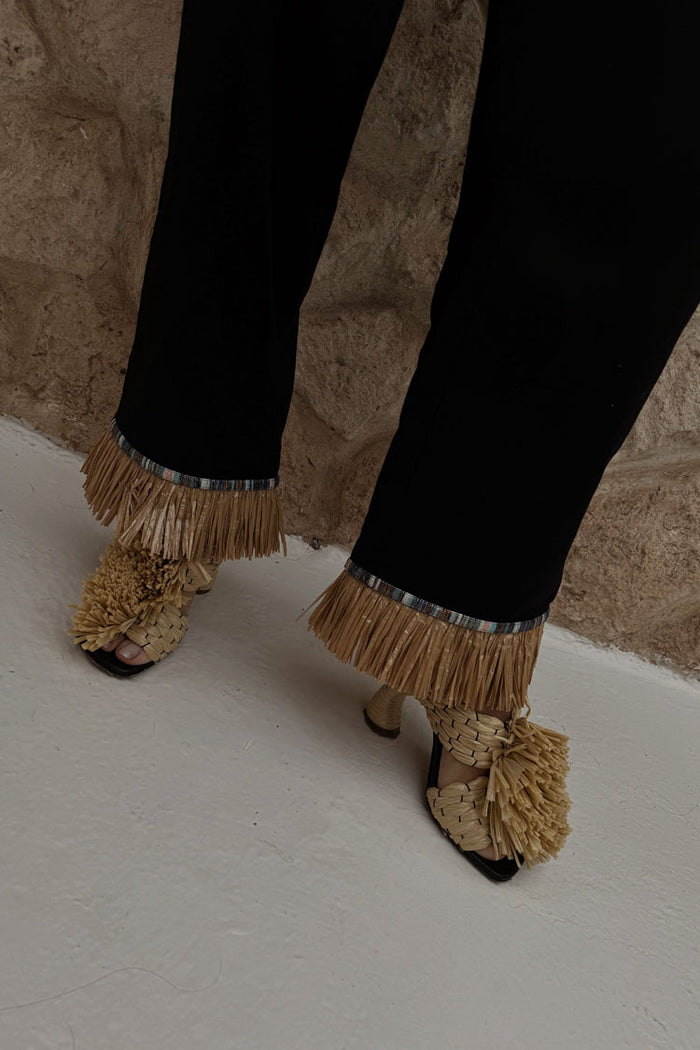 Black Raffia Marra Trousers featuring tan raffia fringe at the hem, styled with woven raffia sandals.