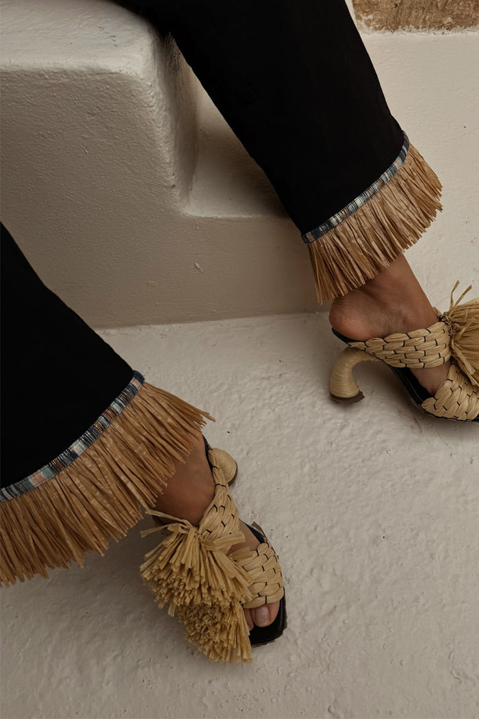 Black Raffia Marra Trousers featuring fringe hems, styled with woven raffia heels on white steps.