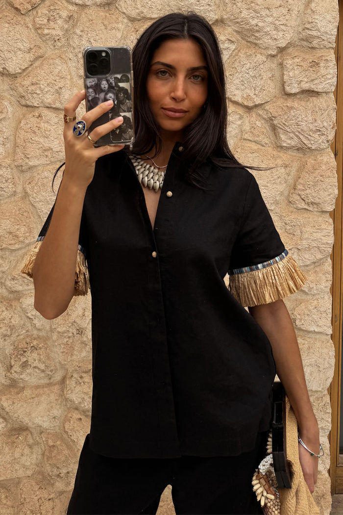 Black Raffia Shirt with raffia fringed sleeves and shell-style buttons, styled in a mirror selfie.