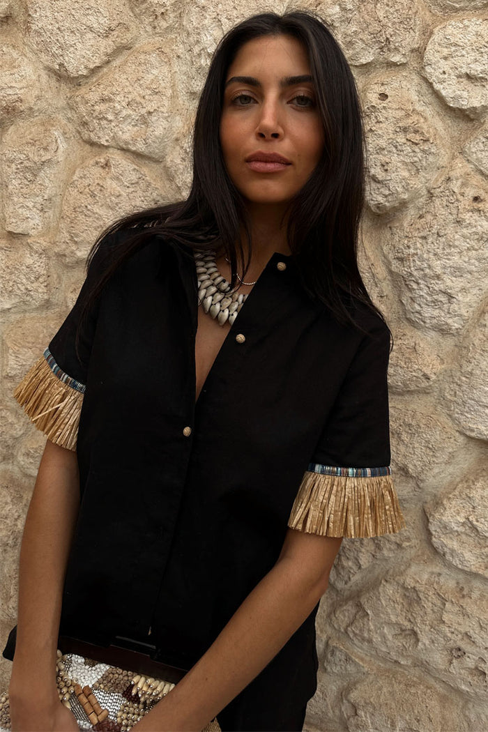Black Raffia Shirt featuring tan raffia fringed sleeves and crochet shell-esque buttons against a stone wall.