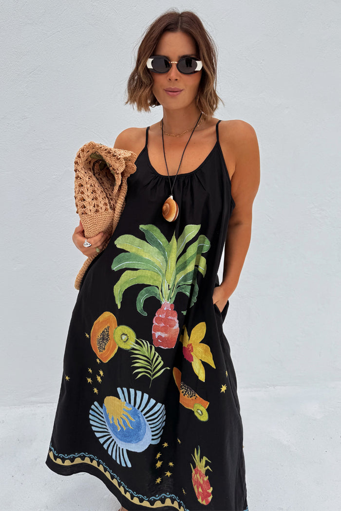 Black Rio Maxi Dress with a vibrant tropical fruit print, styled with a shell necklace and straw hat.