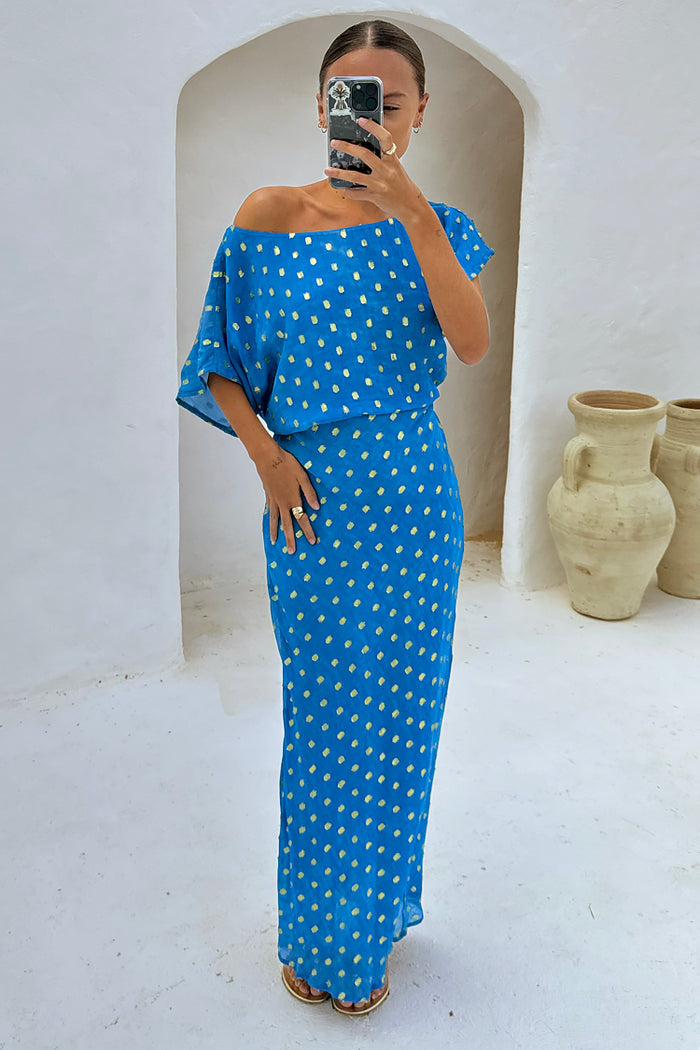 Blue Jacquard Tilly Dress with gold flecks and an off-the-shoulder neckline, styled for a vacation in a white archway.