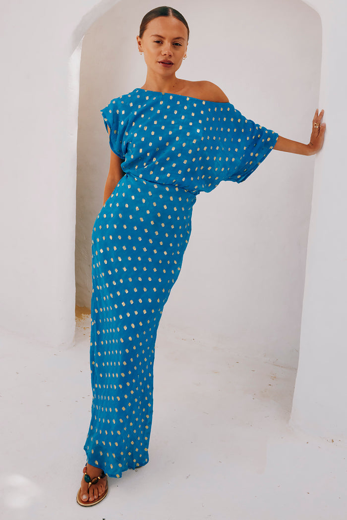 Blue Jacquard Tilly Dress with gold metallic dots and an off-the-shoulder silhouette leaning against a white archway.