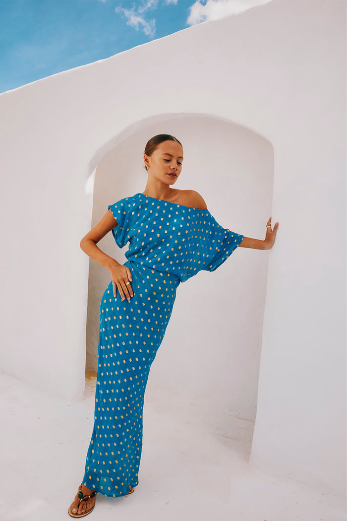 Blue Jacquard Tilly Dress with gold metallic dots and an off-the-shoulder silhouette, posed against a white arch.