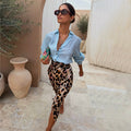 Iconic Brown Leopard Jaspre Skirt in satin, styled with a light blue shirt and sunglasses in an outdoor setting.