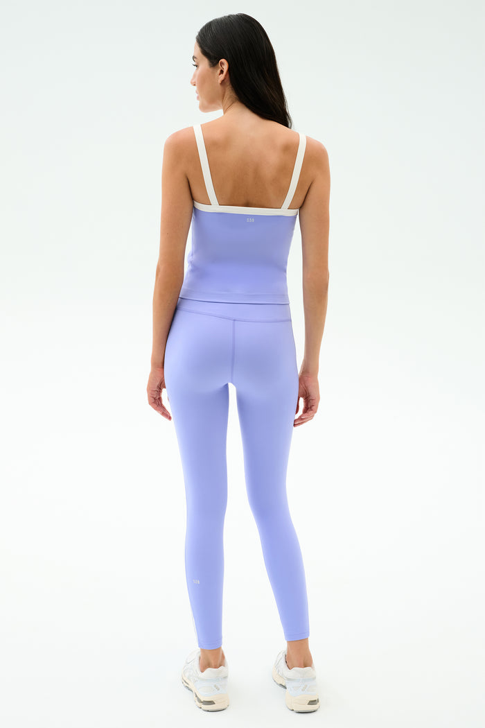A woman faces away on a white background, wearing SPLITS59's Clare High Waist Rigor 7/8 leggings in Purple Haze/White—ideal for the gym—paired with a light purple athletic top and white sneakers.