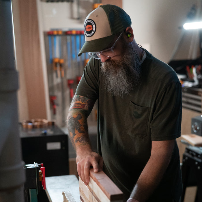Man wearing curved brim work hats with built in pencil holder and BRUNT logo in army green while using table saw * 0