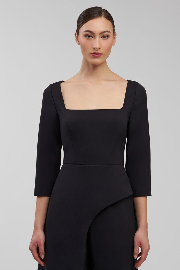 Woman wearing a black walk thru jumpsuit with a square neckline and long sleeves on a gray background