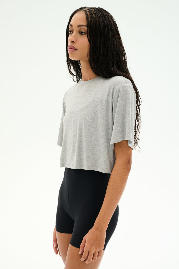 A woman wears the SPLITS59 Djuna Oversized Crop Jersey Tee in Heather Grey, paired with black high-waisted shorts, standing against a plain white background.