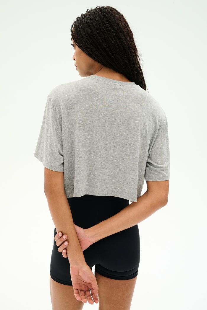 A person with long braided hair stands facing away, wearing the SPLITS59 Djuna Oversized Crop Jersey Tee in Heather Grey and black shorts, hands clasped behind their back against a plain light background.