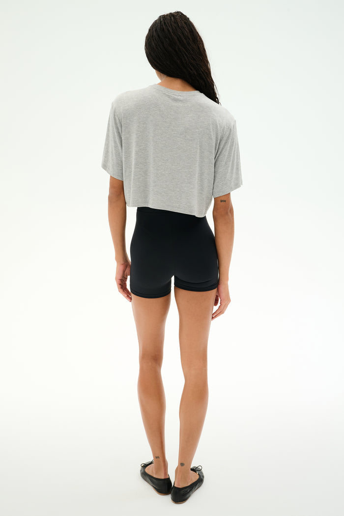A person stands facing away against a white background, wearing SPLITS59's Djuna Oversized Crop Jersey Tee in Heather Grey, paired with black shorts and black flats.