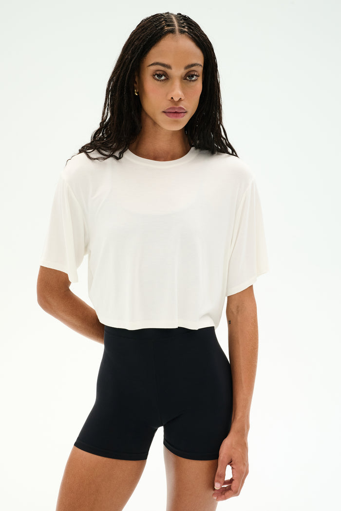 A woman faces forward against a white background, wearing the SPLITS59 Djuna Oversized Crop Jersey Tee in white paired with high-waisted black stretchy shorts.