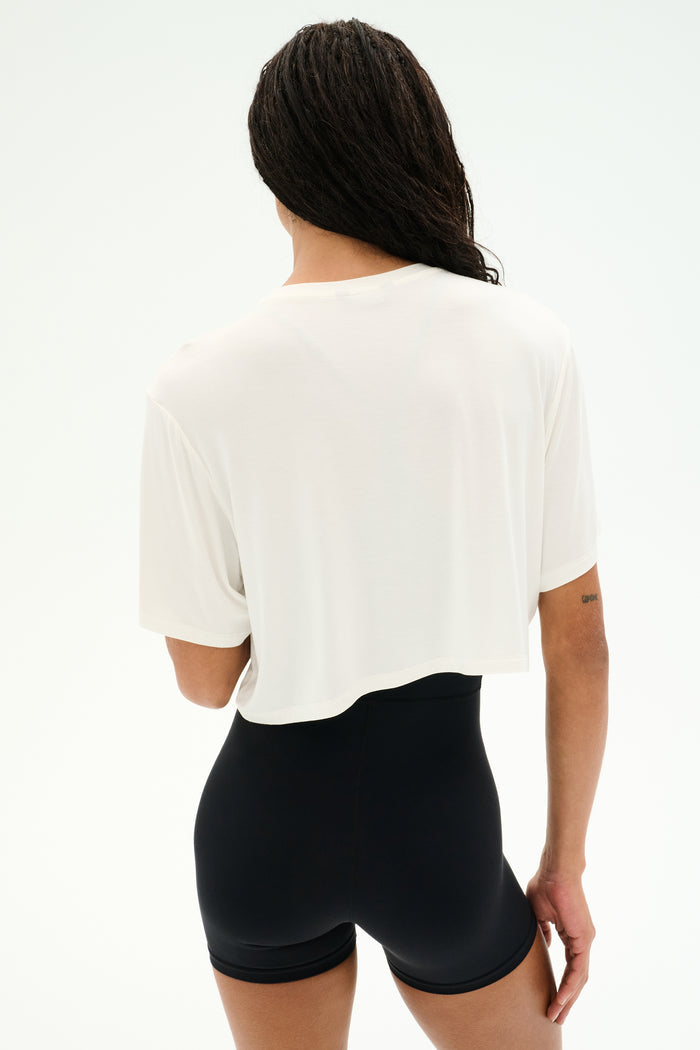 A person with long braided hair wears the SPLITS59 Djuna Oversized Crop Jersey Tee in white, paired with black high-waisted shorts, standing with their back to the camera against a plain light background.