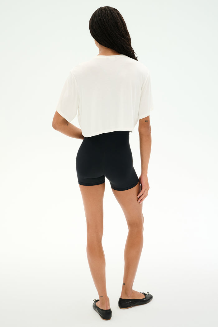 A person stands on a white background, facing away, wearing the SPLITS59 Djuna Oversized Crop Jersey Tee in white, paired with black spandex shorts and black flats.