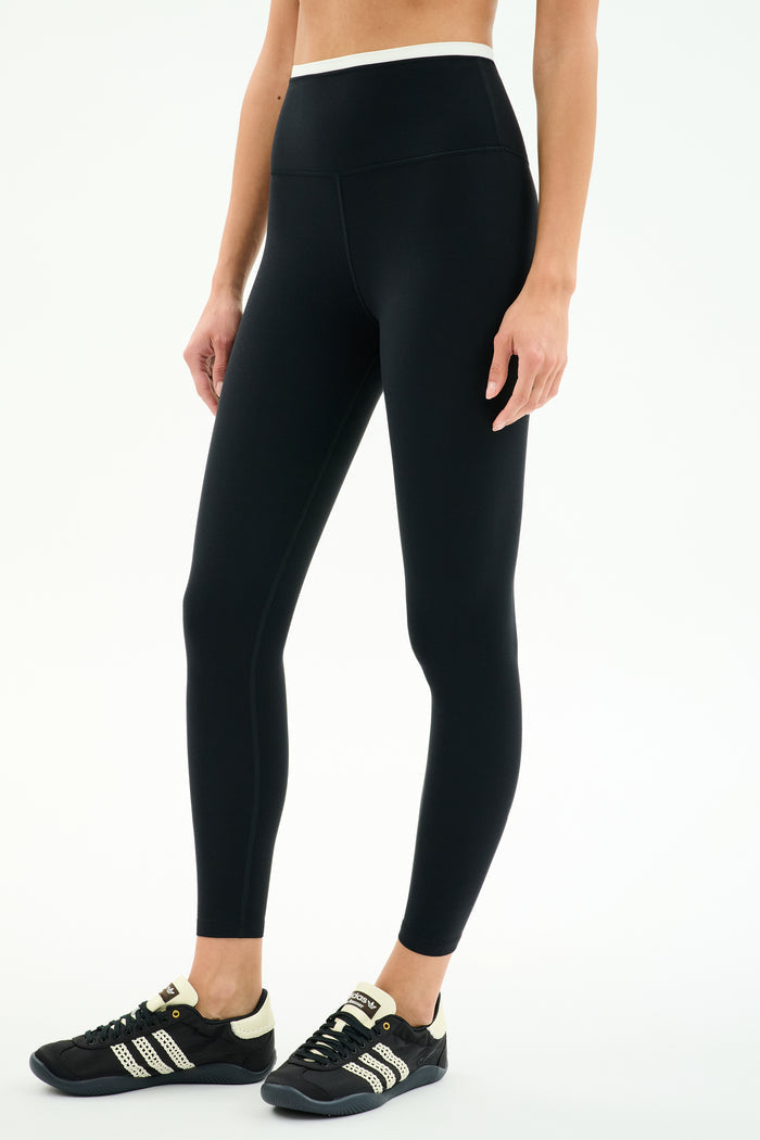 A person wearing SPLITS59 Dual High Waist Airweight 7/8 leggings in Black/White and black athletic shoes with white stripes, standing against a plain white background.