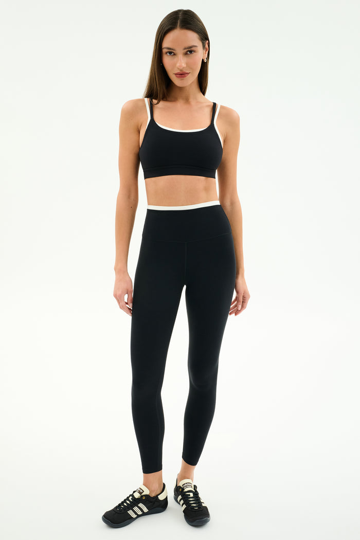 A woman faces forward against a plain white background wearing the SPLITS59 Dual High Waist Airweight 7/8 leggings in Black/White, paired with a black sports bra and black athletic shoes.