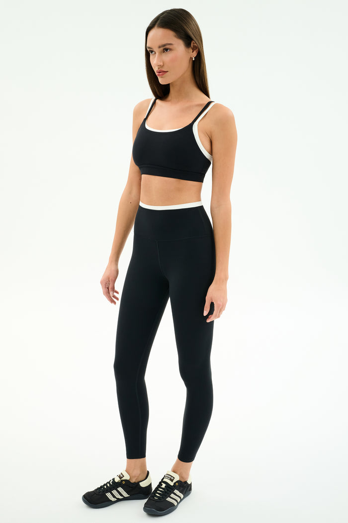 A woman stands against a white background wearing the SPLITS59 Dual High Waist Airweight 7/8 leggings in Black/White, paired with a black sports bra and black Adidas sneakers.